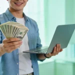 Beginner investor holding cash in one hand and a piggy bank with a laptop in the other, symbolizing investing in yourself versus the stock market”