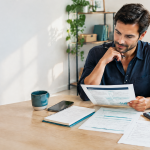 Solo male business owner reviewing a business plan, customer notes, startup costs, and cash-flow paperwork at a desk.